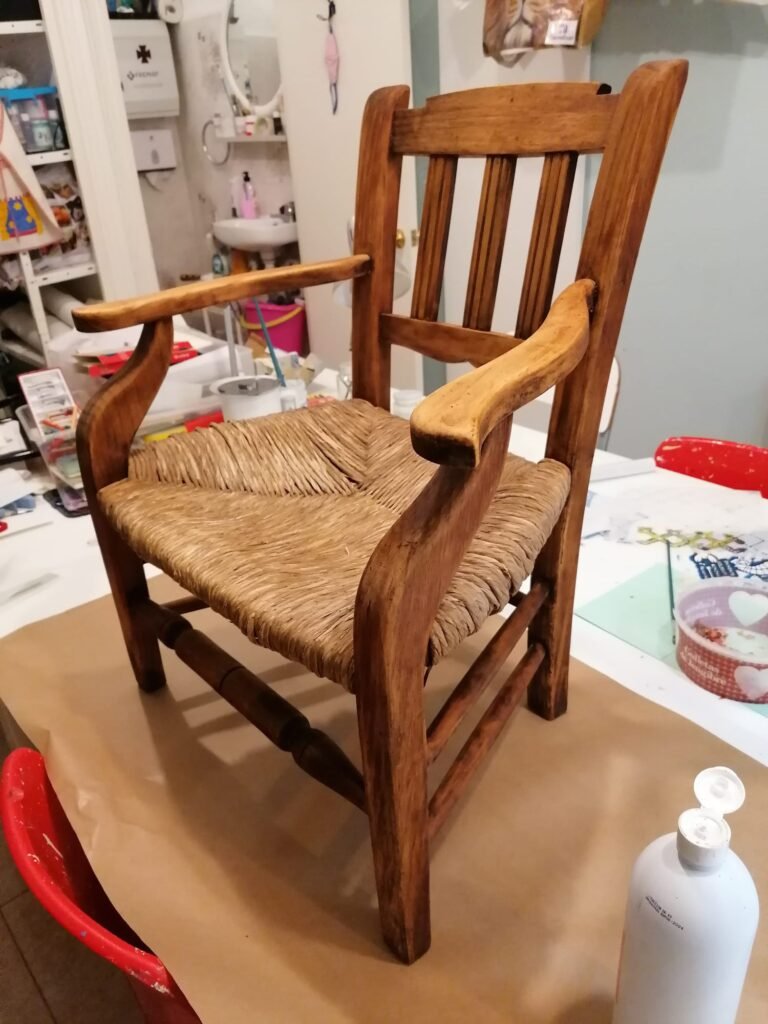 Silla de madera con asiento de cuerda tejida, colocada sobre una mesa. La silla tiene reposabrazos curvos y un respaldo de listones verticales.